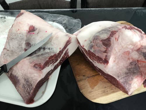 El cliente muestra dos piezas de pecho crudo antes de prepararlas, una sobre una bandeja y otra en una tabla para cortar.