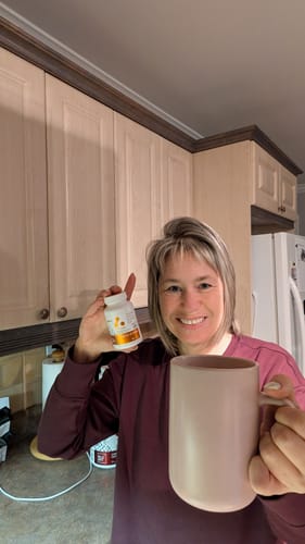 Cliente souriante tenant la bouteille de Vitamine D3 + K2 d'ATP Lab et une tasse dans sa cuisine.