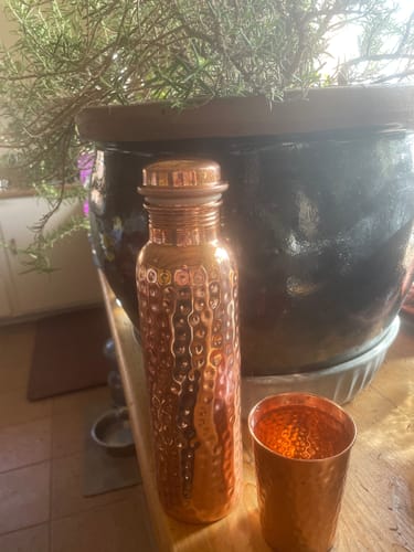 Customer's hand-hammered Pure Copper Water Bottle and matching cup sitting on a wooden surface next to a potted plant.