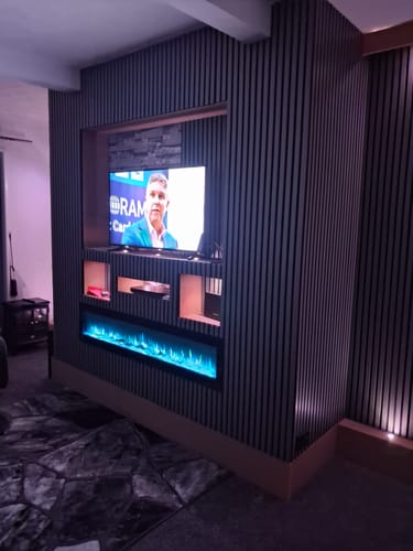Customer's dark Acoustic Slatted Wall Panels installed as a feature media wall around a TV and electric fireplace.