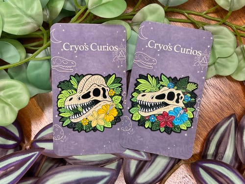 Customer's two custom enamel pins of dinosaur skulls with tropical flowers, displayed among plant leaves.
