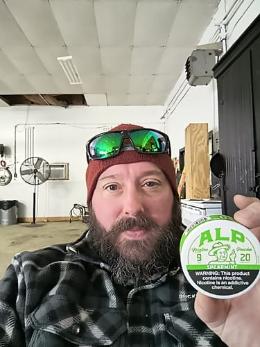 A reviewer holds up a round tin of ALP Spearmint Nicotine Pouches in a garage.