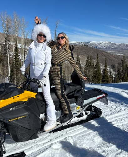 Two customers on a snowmobile, one in a white one-piece ski suit and the other in a houndstooth version.