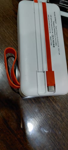 Reviewer's white Powerbank KPB037 with its built-in orange USB-C cable and matching wrist strap on a wooden surface.
