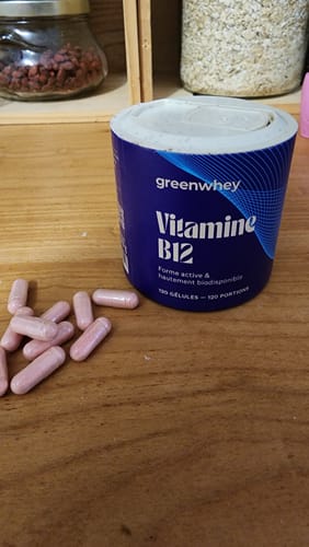 Client montrant le pot de Vitamine B12 bleu à côté de gélules roses sur une table en bois.
