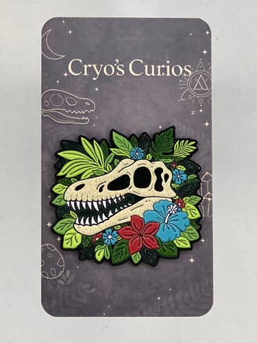 Customer's sparkly custom enamel pin of a dinosaur skull with red and blue flowers, on its backing card.