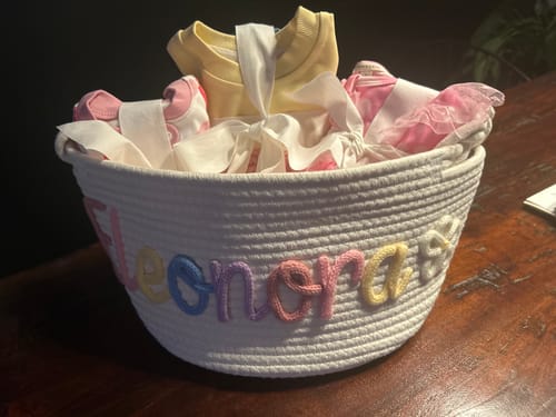Customer's personalized white knitted basket with 'Eleonora' in multicolor yarn, filled with baby clothes on a wooden table.