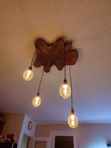 Ein Kunde zeigt die montierte NATURA Baumscheibe Deckenlampe, die mit vier Glühbirnen einen Raum warm beleuchtet.