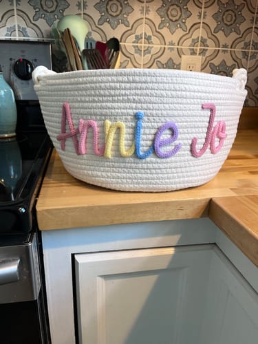 Customer's white personalized knitted basket with 'Annie Jo' written in multicolor yarn on a counter.