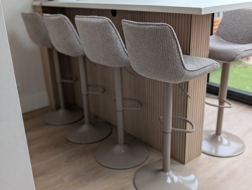 Recensent toont vier zandkleurige WOOOD Marten barstoelen die netjes aan een kookeiland staan.