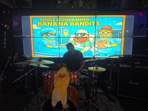 Reviewer at a drum kit on stage, with the band's custom artwork for the drumhead skin displayed on a large screen.