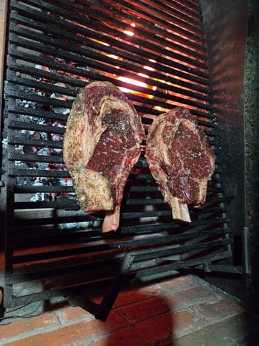 Cliente asando dos Rib Eye Cowboy Steaks sazonados en una parrilla sobre brasas.