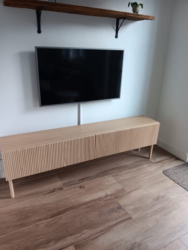 Klant toont het lichte, geribbelde TV-meubel Sian in een woonkamer onder een aan de muur gemonteerde televisie.