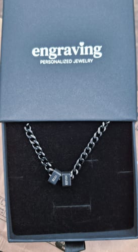Reviewer's Legacy necklace in its black gift box. The dark chain has two beads engraved with the names 'Ella' and 'Kevin'.