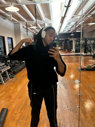Reviewer in a gym wearing the black Megàlo 3/5 Sleeve Hoodie with the hood up over headphones while taking a mirror selfie.