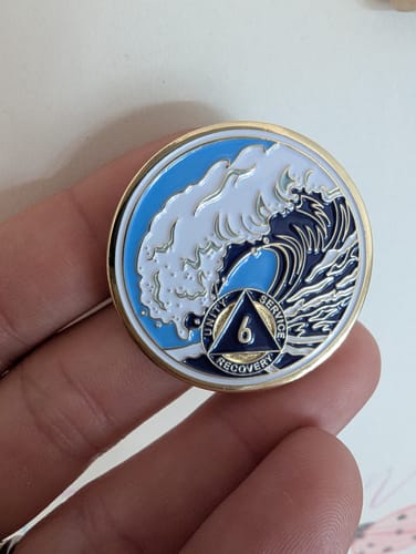 Customer holds the front of the 6-year crashing wave sobriety medallion, showing the detailed blue and white enamel design.