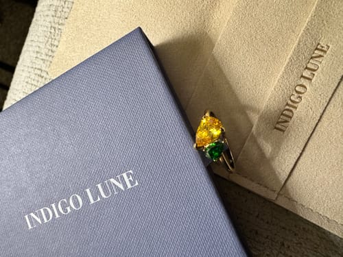 Customer's Leesha Birthstone Ring with yellow and green stones, displayed with its blue box and beige pouch.