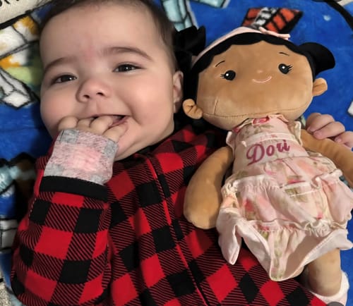 A customer's baby in a red plaid onesie smiles while lying next to a Personalized Princess Petal doll with 'Dori' on the dress.