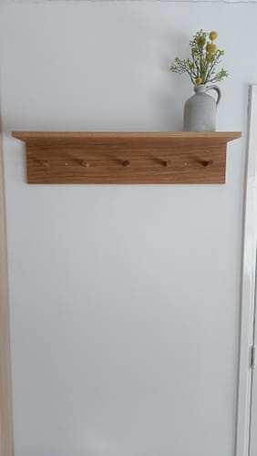 Recensent toont de eikenhouten Ferm Living Place wandkapstok, opgehangen aan een witte muur met een vaasje erop.