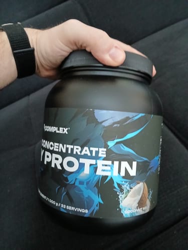 Reviewer holding a large black tub of Complex CFM Whey Protein in the Coconut Milk flavor.