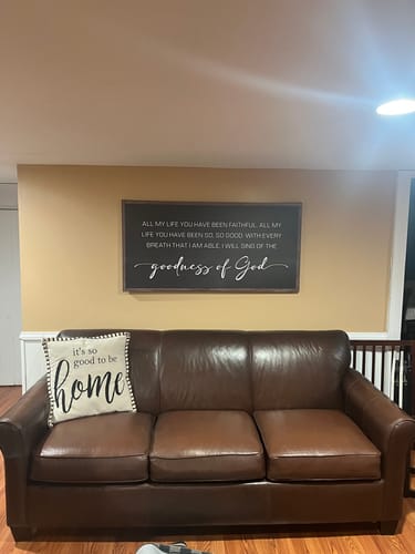 Customer's Goodness Of God Faith Sign II hanging on a wall above a brown leather sofa.