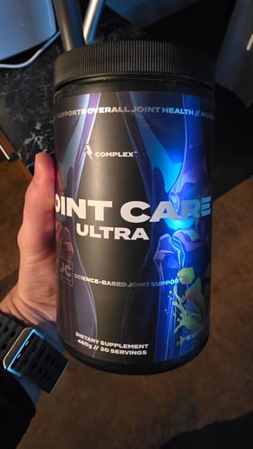 A reviewer's hand holding a large black tub of Complex Joint Care Ultra supplement.