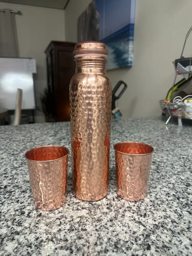 Reviewer's hand-hammered Pure Copper Water Bottle with two matching cups sitting on a granite countertop.