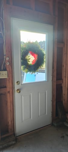 Customer's installed white Steel Entry Door Slab with a half lite glass panel, seen from an unfinished interior.