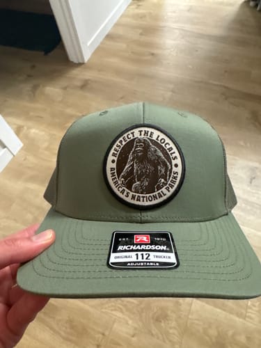 Customer holding the sage green Respect The Locals Trucker Hat, showing the Bigfoot patch.