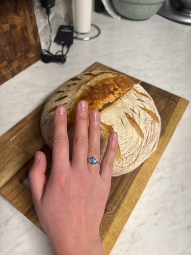 Customer's hand wearing the Leesha Birthstone Ring with two light-blue stones on a gold band, resting on a loaf of bread.