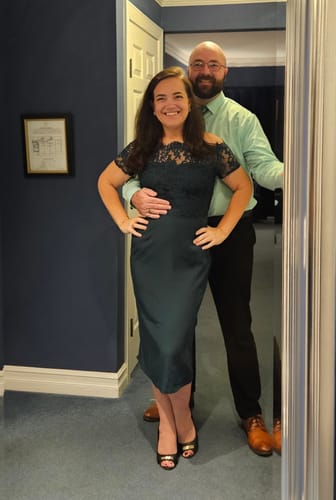 Customer smiling while posing in the dark green lace off-shoulder dress in a hallway.