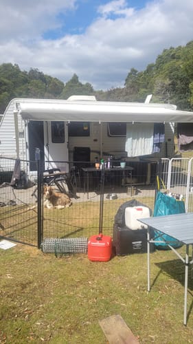 A wide shot of the 8 Panel Pet Dog Playpen set up next to a caravan, with a dog resting inside.