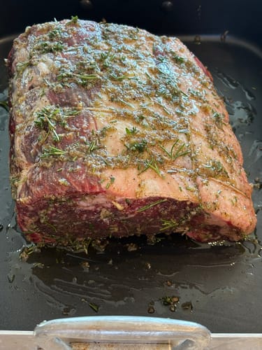 Customer's raw Prime Rib Roast generously seasoned with herbs and spices in a roasting pan.