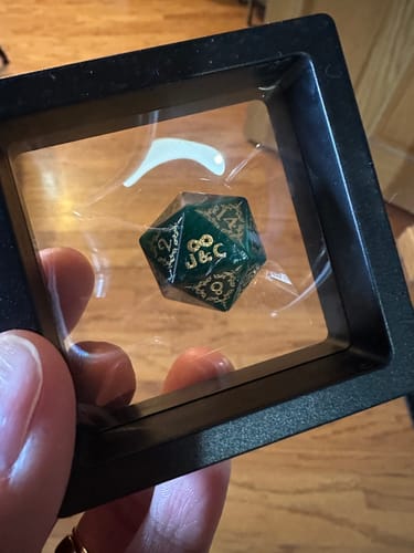 Customer holding a custom green die in a display case, showing gold initials 'J&C' and an infinity symbol.