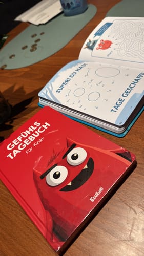 Kunde zeigt das rote Gefühlstagebuch für Kinder geschlossen und daneben offen mit einer Rätselseite.