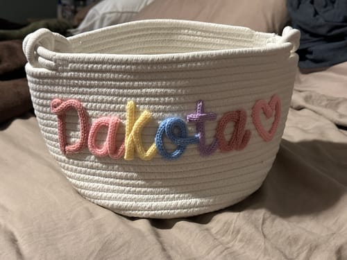Customer's personalized knitted basket in white, with the name 'Dakota' in multicolor yarn and a pink heart icon.