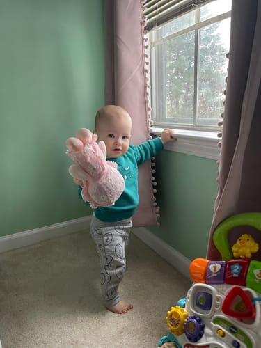 Reviewer's baby standing in a room and holding the soft pink Personalized Daisy Baby Girl doll.