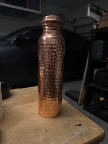 Customer's hand-hammered Pure Copper Water Bottle standing on a wooden table in a garage.