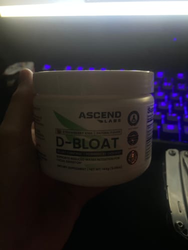 Customer holding a white tub of D-BLOAT Strawberry Kiwi powder in a dimly lit room.