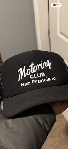 Reviewer holding a black trucker hat with custom white embroidery reading 'Motoring CLUB San Francisco'