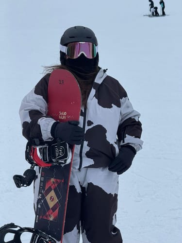Close-up of a reviewer on a slope wearing baggy off-white snow pants with a dark pattern, holding a snowboard.