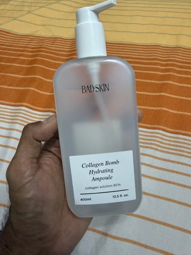Reviewer holding the translucent 400ml bottle of Bad Skin Collagen Bomb Hydrating Ampoule against a striped background.