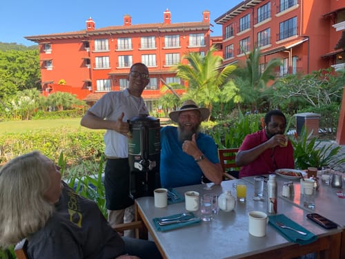 Reviewer giving a thumbs-up as a staff member serves coffee from an urn at a sunny outdoor breakfast.