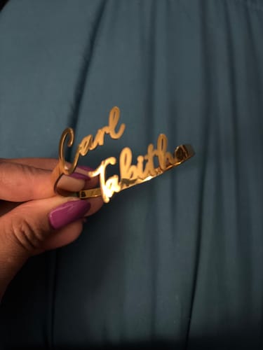 Customer holds their gold Personalized Two-Name Bangle with the names 'Carl' and 'Tabitha' against a teal background.
