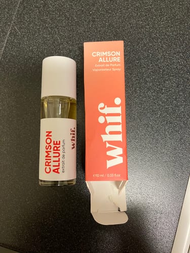 Reviewer's small bottle of Crimson Allure perfume shown next to its reddish box on a dark surface.