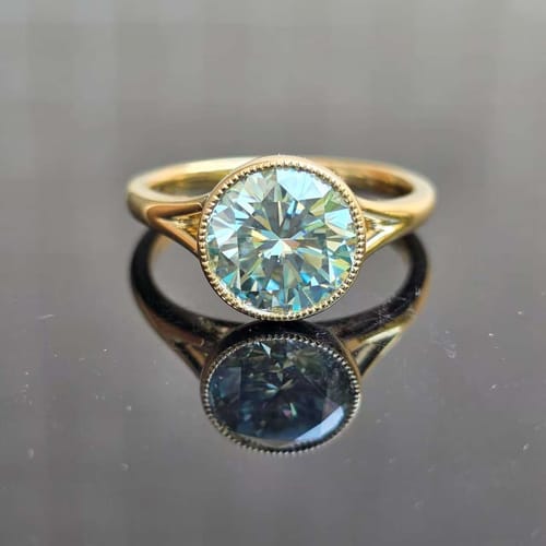 Customer's bezel solitaire ring with a sparkly bluish-green stone in a gold band, seen from above on a reflective surface.
