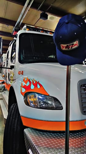 Customer's blue Snapback Hose Hat displayed on a stand in front of a white fire truck in a station.