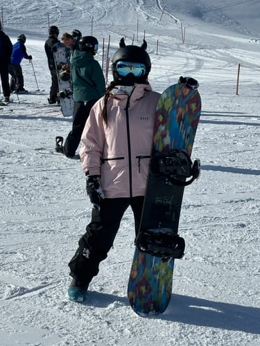 Reviewer on a snowy slope wearing the baggy black snow pants with a pink jacket and holding a snowboard.