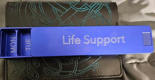Customer's blue Weekly Case, engraved with 'Life Support', is partially open, revealing compartments for Monday and Tuesday.