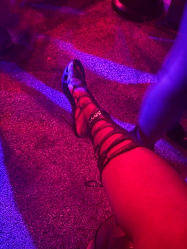 Reviewer wearing the strappy black Snake Heel on a dark floor with dramatic red and purple lighting.